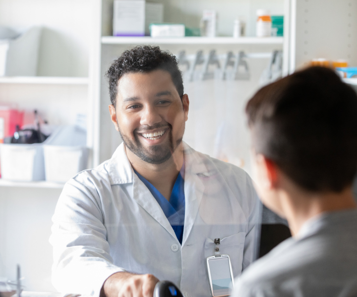 A smiling male pharmacist with short curly hair and a beard is standing behind a counter in a pharmacy, talking to a customer. Shelves with medication and pharmacy supplies are visible in the background.