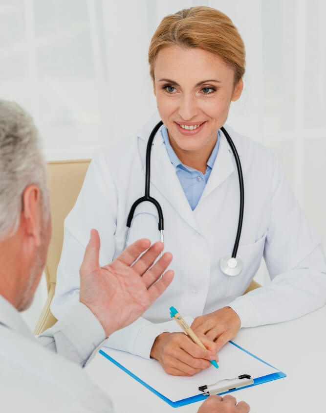 A doctor with a stethoscope around her neck sits at a desk, smiling and holding a pen. As she listens to the patient gesturing beside the blue clipboard, she considers using HIPAA-compliant texting for secure communication.