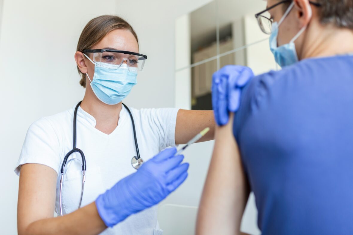 A healthcare professional wearing a mask, safety glasses, and gloves is administering a vaccine to an individual in a blue shirt. The person receiving the vaccine is also wearing a mask. Both are in a clinical setting.