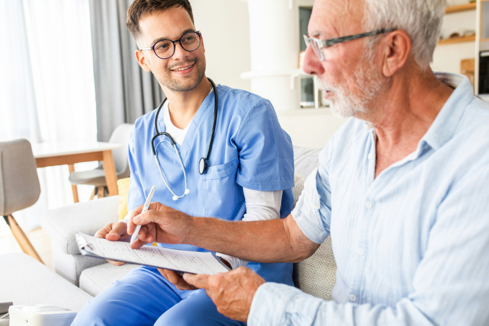 A young healthcare professional in blue scrubs and a stethoscope, smiles while conversing with an elderly man holding a clipboard with papers. They are seated in a well-lit room with furniture in the background. Both appear engaged in a friendly discussion.