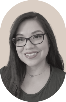 A woman with long dark hair and glasses smiles while looking at the camera. She is wearing a necklace and a dark-colored top. The background is plain and slightly blurred, emphasizing her face.