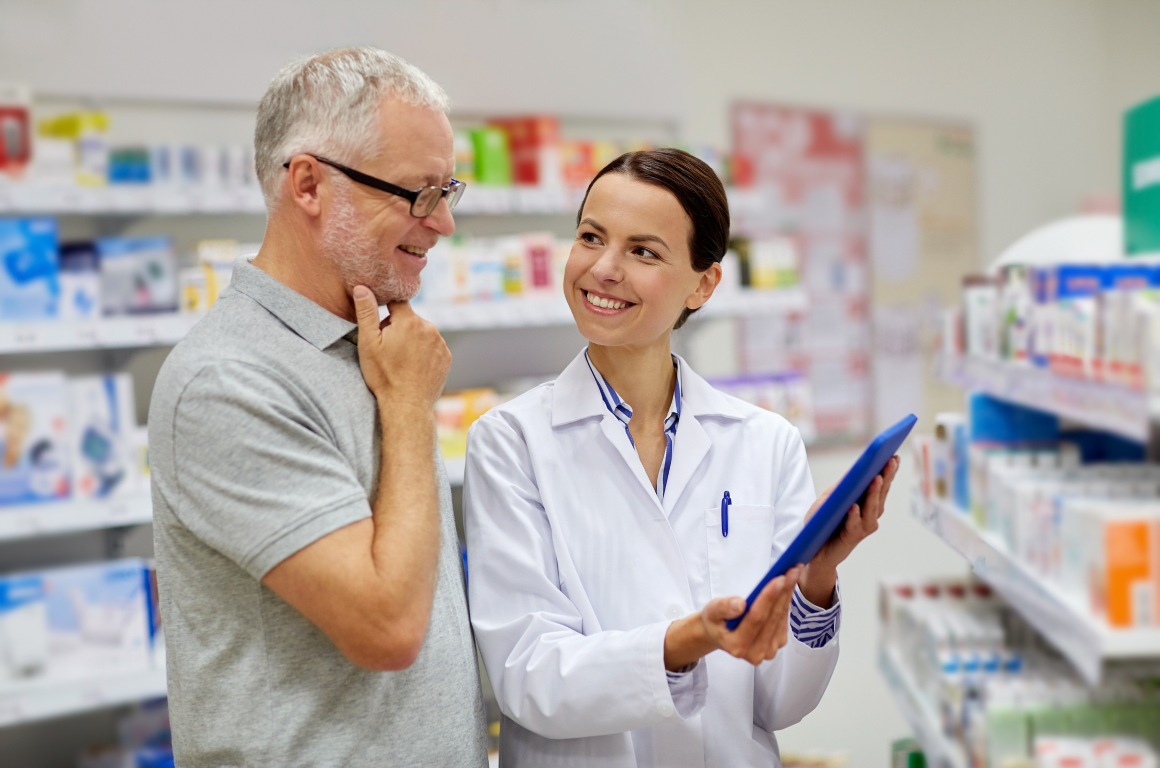 A pharmacist in a white coat smiles while showing a clipboard to an older man in a pharmacy. The man, dressed in a gray polo shirt and glasses, looks attentively at the clipboard. They are surrounded by shelves stocked with various products.