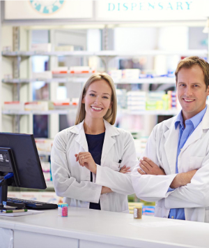 Two smiling pharmacists in white lab coats stand behind a counter in a well-lit pharmacy. Shelves stocked with various medicines and products can be seen in the background. They appear professional and approachable, ready to assist customers.