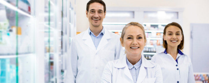 Three smiling healthcare professionals, two women and one man, stand in a brightly lit pharmacy. They are wearing white lab coats, suggesting they are pharmacists or pharmaceutical staff. Shelves filled with products are visible in the background.