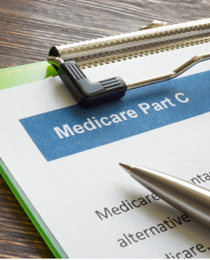 Close-up of a document titled "Medicare Part C" placed on a clipboard with a green cover. A silver pen rests on the paper next to the text. The background surface is a wooden table.