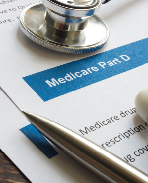 A document titled "Medicare Part D" is partially visible, with some text about Medicare drug coverage. A silver pen, a stethoscope, and white pills are placed on top of the document. The stethoscope's diaphragm is prominently seen, suggesting a healthcare context.