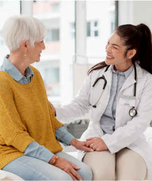 A doctor with a stethoscope around her neck is sitting and smiling warmly at an elderly woman with short white hair who is wearing a mustard yellow sweater. The doctor has her hand on the woman's arm. They appear to be in a medical office.