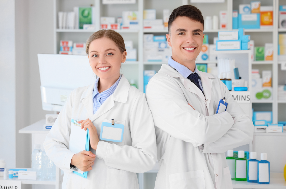 Two young pharmacists in lab coats smile confidently in a pharmacy setting. Shelves filled with medicine and health products are visible in the background. One holds a clipboard, and they both exude professionalism.