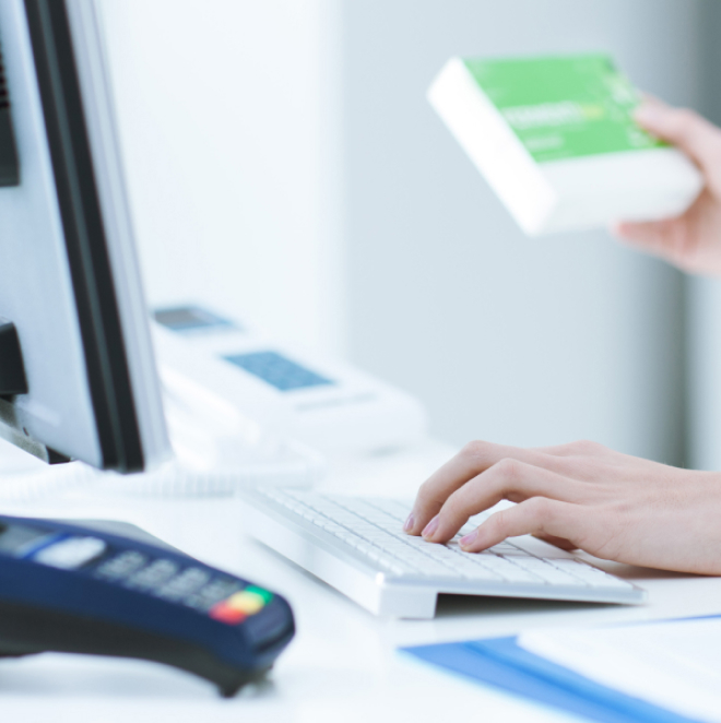 Hands typing on a white keyboard next to a monitor. A person holds a green and white box in the background. A payment terminal is visible in the foreground on a white desk.