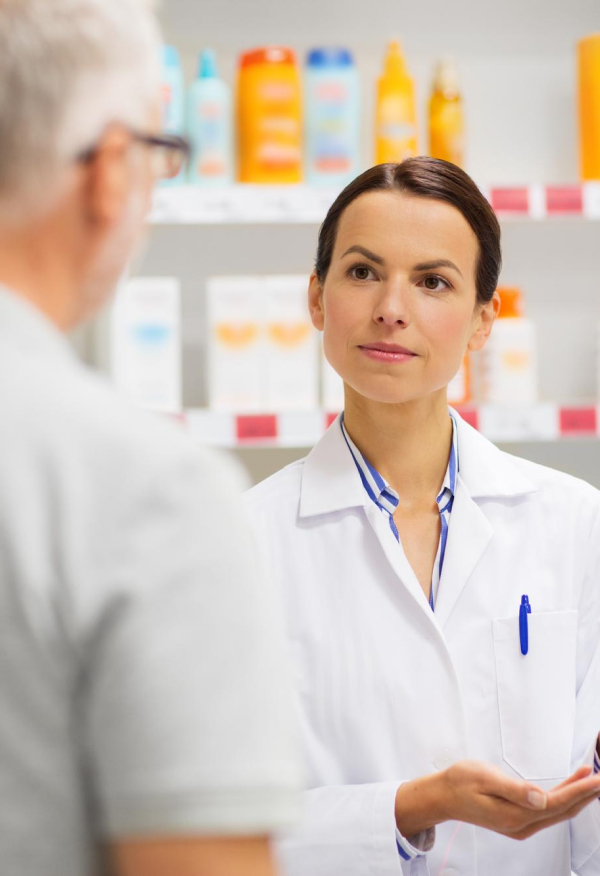 A female pharmacist in a white coat listens attentively to a customer in a pharmacy. Shelves filled with various products are visible in the background. The pharmacist holds a medication package while engaging in conversation.