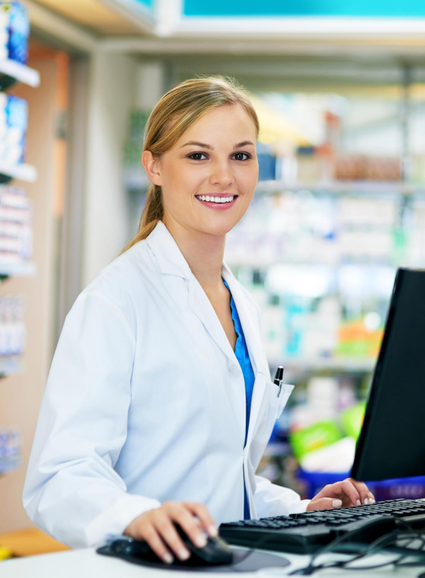 A pharmacist in a white coat is smiling at the camera while working on a computer in a pharmacy. Shelves with various products are visible in the background.