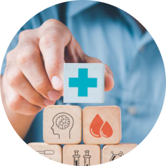 A person in a blue shirt places a block with a blue medical cross atop a stack of wooden blocks. The other blocks display icons of a brain, blood droplets, and medical supplies, representing health and healthcare concepts.