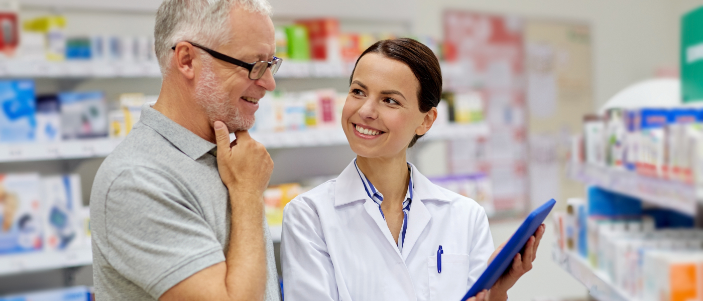 A pharmacist in a white coat holds a blue clipboard and smiles while talking to a man with glasses and a gray shirt in a pharmacy. Shelves with various products are visible in the background.