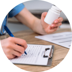 A person in medical attire holds a white pill bottle in one hand while writing on a clipboard with the other, sitting at a wooden desk. The focus is on the hands, with medical documents visible in the background.