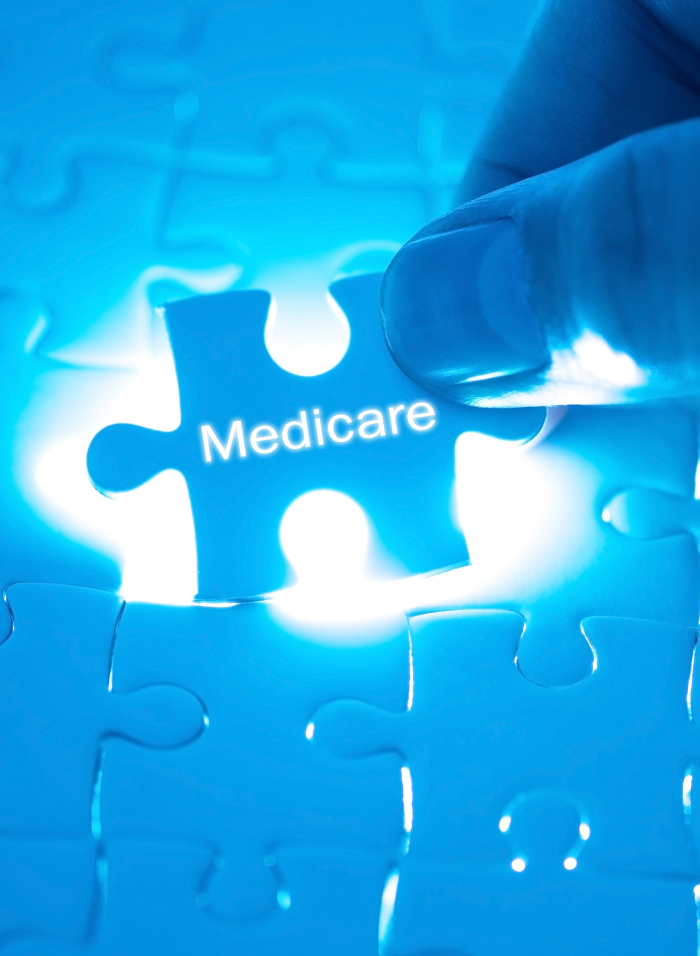 Close-up of a hand fitting a glowing puzzle piece labeled "Medicare" into a puzzle. The puzzle pieces are illuminated in blue light, creating a bright and focused image centered on the Medicare piece.