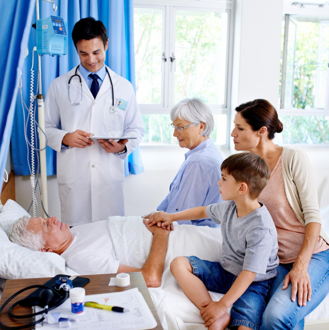 A doctor stands by the bedside of an elderly patient in a hospital room. A young woman sits next to the patient, holding hands, with a child on her lap. An older woman sits nearby, watching the scene. Medical equipment is visible.