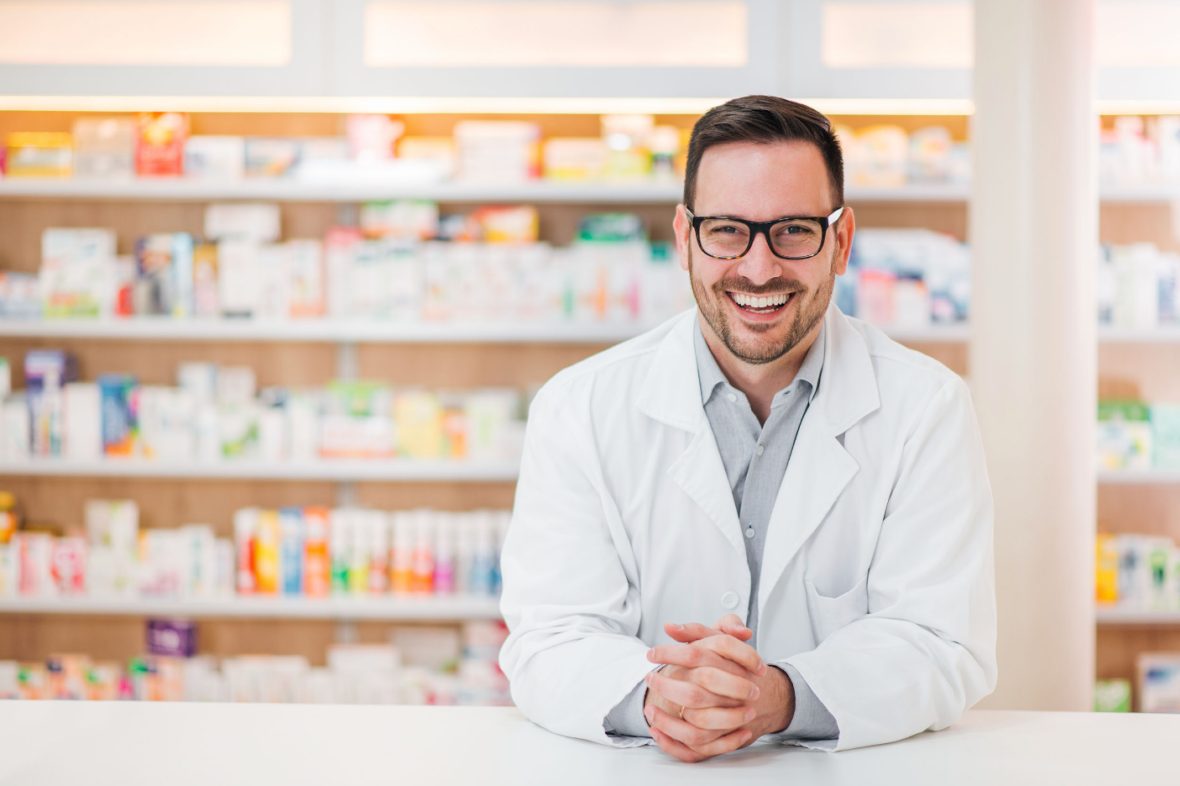 A smiling pharmacist stands in front of shelves filled with various medications and products. He is wearing a white lab coat and glasses, with his hands clasped on the countertop.