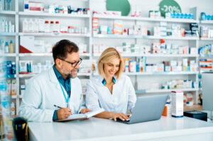 Two pharmacists in white coats are working together in a pharmacy. They are standing at a counter; one is taking notes while the other is using a laptop. Shelves filled with various medicines and products are visible in the background.