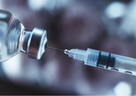 A syringe needle is being inserted into a vial, ready for drawing liquid. The background is softly blurred, emphasizing the focus on the medical instruments, suggesting preparation for an injection or medical procedure.