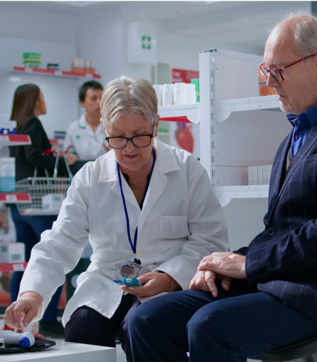 A pharmacist in a white coat is seated next to an elderly man, showing him how to use a medical device. Shelves with medicine and other staff members are visible in the background.