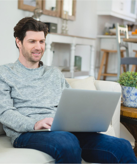 A man with dark hair is sitting on a white sofa using a laptop. He's wearing a gray sweater and blue jeans. The background shows a modern, bright living room with a plant and some furniture.