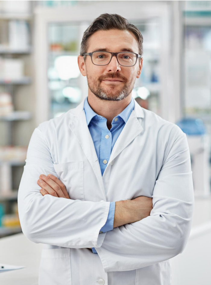 A male doctor with short hair and glasses stands confidently with arms crossed. He wears a white lab coat over a blue shirt. The background shows a bright, blurred medical or pharmacy setting with shelves.