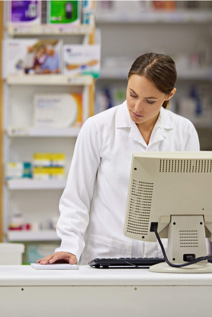 A pharmacist wearing a white lab coat stands behind a pharmacy counter, looking at a computer monitor. Shelves with various medicines and health products are visible in the background.