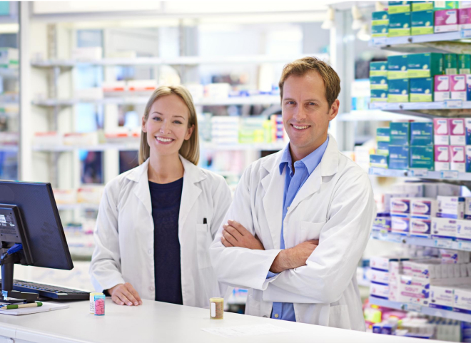 Two pharmacists in white coats stand behind a counter in a pharmacy. They smile at the camera, with shelves filled with medicine boxes in the background. A computer and medication bottles are on the counter.