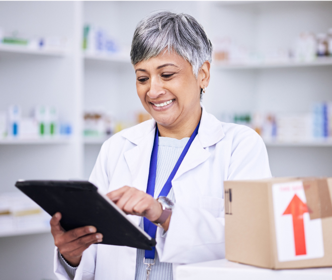 A pharmacist with short gray hair smiles while using a tablet. She wears a white lab coat and stands in a pharmacy with shelves of medication in the background. A cardboard box with a red arrow is on a counter nearby.