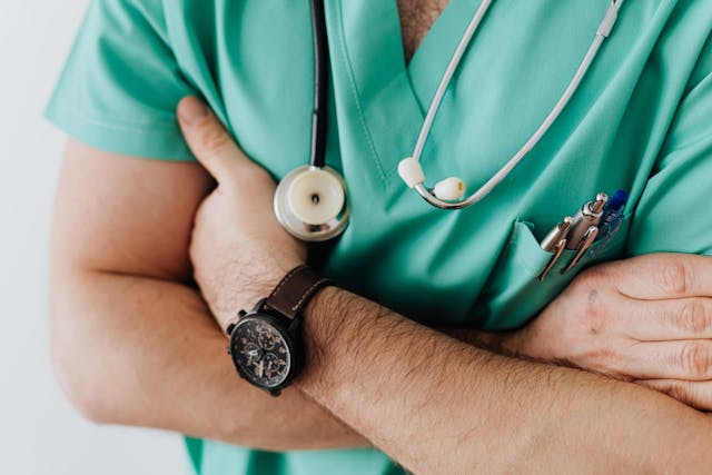 A person in green medical scrubs with a stethoscope around their neck is crossing their arms. A watch is visible on their wrist, and pens are tucked into the chest pocket. The background is a plain, light color.