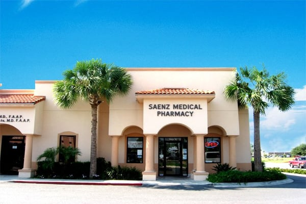 A pharmacy building with a sign reading "Saenz Medical Pharmacy" on the front. It has a beige exterior, arched windows, and palm trees in front. The sky is clear and blue.