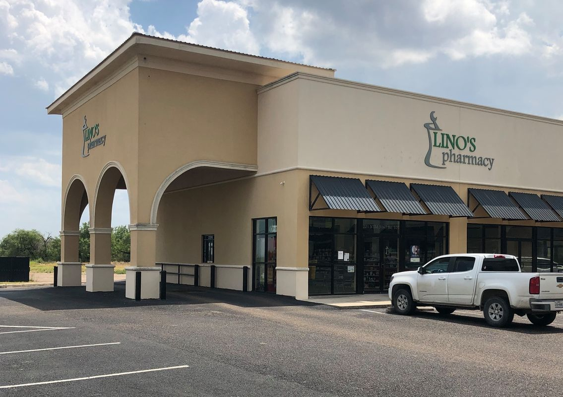 A single-story building with a sign that reads "Lino's Pharmacy" on the facade. The structure is tan with black awnings over windows. A white pickup truck is parked in front. The sky is partly cloudy.