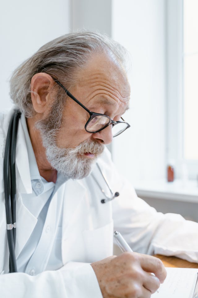 Elderly doctor with gray hair and glasses, wearing a white coat and stethoscope, writing on a notepad at a desk next to a bright window.