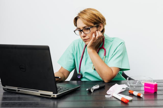 A person wearing green scrubs and a stethoscope sits at a desk, looking at a laptop with a contemplative expression. Nearby are pens, a mask, protective goggles, and a small pink object, all on a dark wooden table.