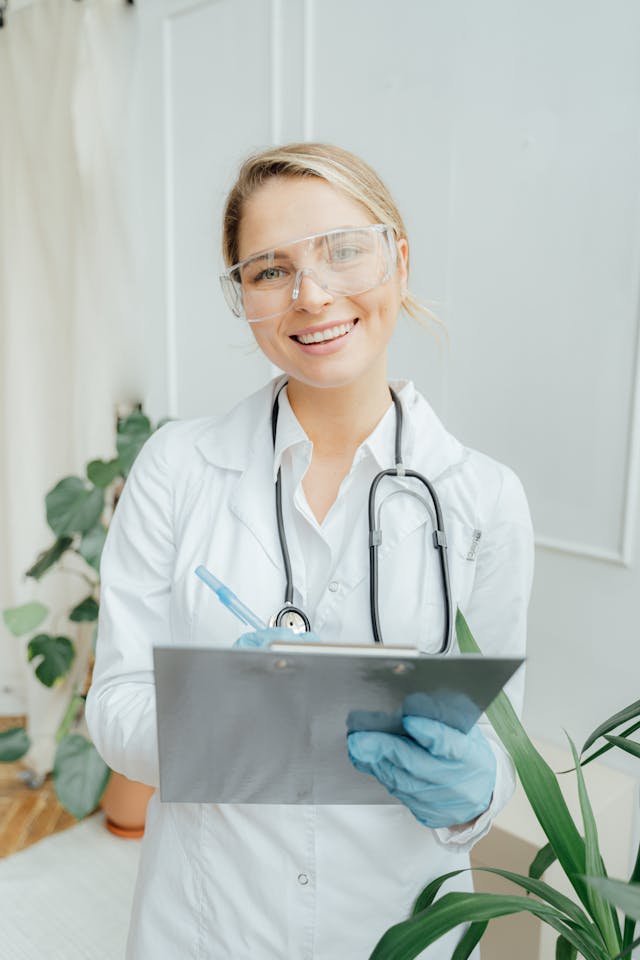 Smiling healthcare professional in a white coat and protective glasses, holding a clipboard with a pen. A stethoscope hangs around their neck. They are indoors, with plants visible in the background.