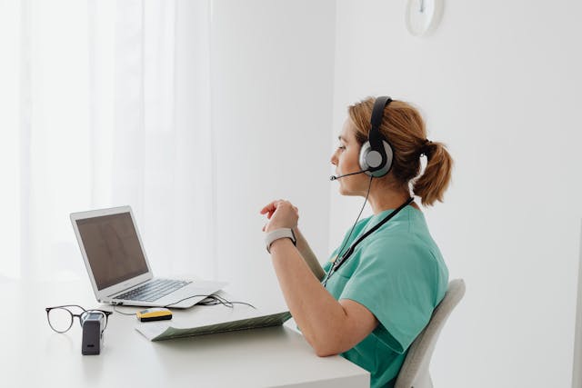 A healthcare professional in green scrubs sits at a white desk, using a laptop and wearing a headset. Glasses and a small device are on the desk. The setting is bright and minimalistic, with a wall clock in the background.