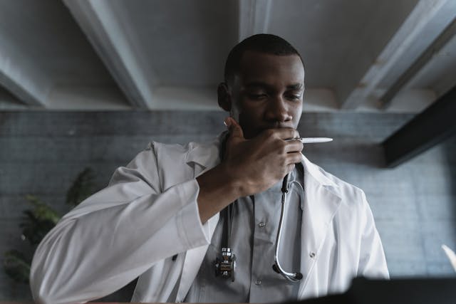 A doctor in a white coat, with a stethoscope around his neck, holds a pen near his mouth. He appears deep in thought, looking down at something off-screen. The background features a concrete wall and ceiling beams.