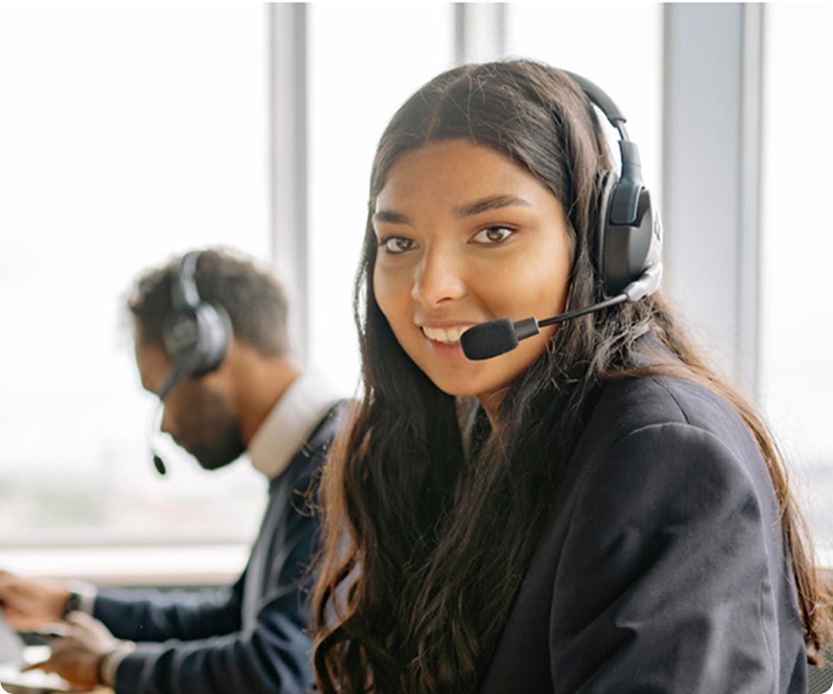 A woman with long hair, wearing a headset and a blazer, smiles at the camera. She is sitting at a desk, with a blurred man in the background also wearing a headset and working at a computer. Large windows are in the background.