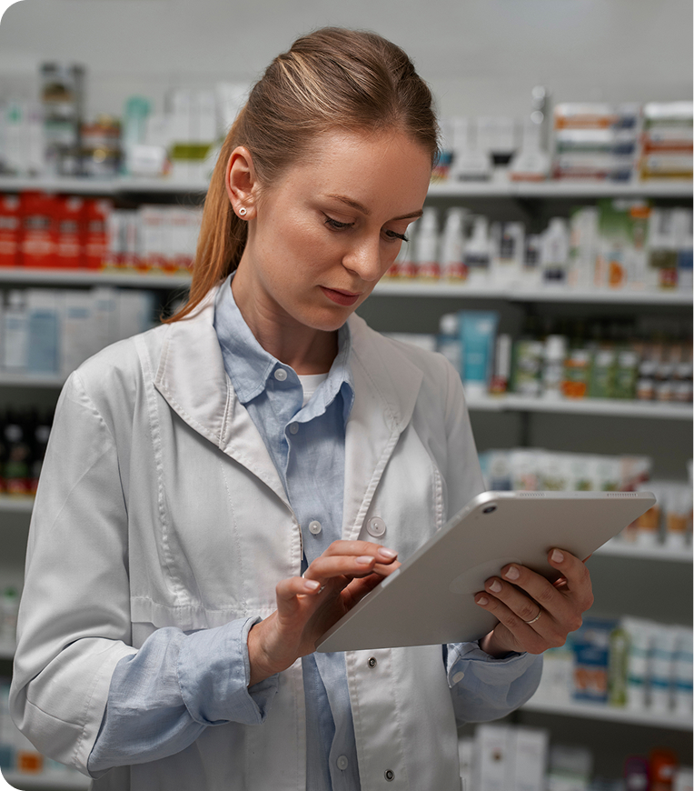 A pharmacist in a white coat stands in a pharmacy, using a tablet. Shelves filled with various medicines and products are visible in the background. She focuses intently on the screen while using her right hand to navigate.