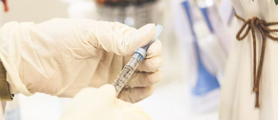 A gloved hand holds a syringe with a blue cap. The syringe is filled with liquid, and the background shows out-of-focus medical equipment.