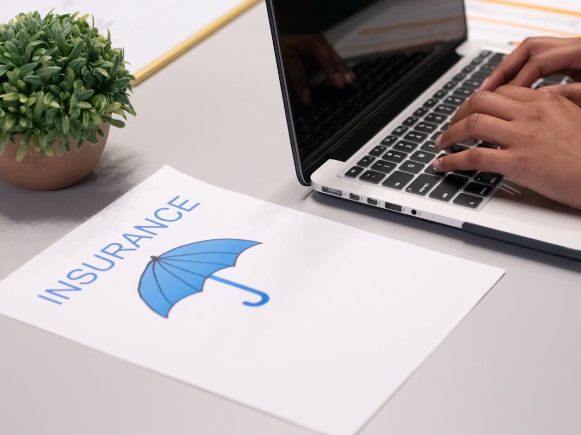 A person types on a laptop next to a document featuring a blue umbrella and the word "INSURANCE." A small potted plant is visible beside the laptop on the desk.