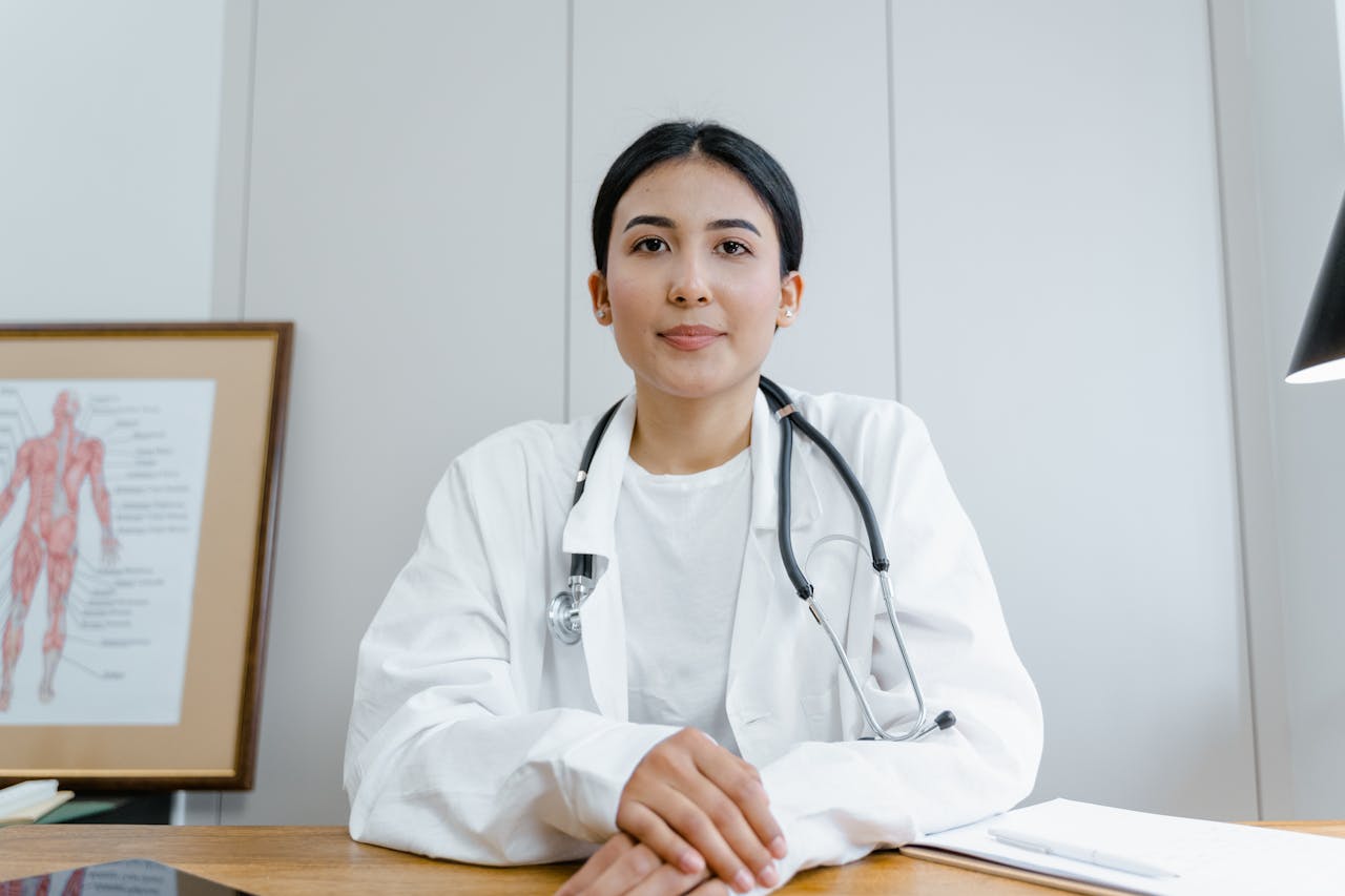 A doctor with a stethoscope around her neck sits at a desk, wearing a white coat. There's a diagram of the human body in the background, and she is in a well-lit room, appearing professional and focused.