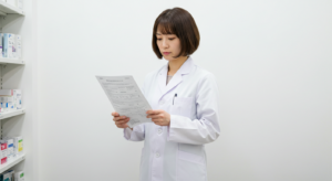 A person wearing a white lab coat reads a sheet of paper in a room with shelves stocked with various boxes. The background is plain white, and the shelves contain organized medical or pharmaceutical supplies.