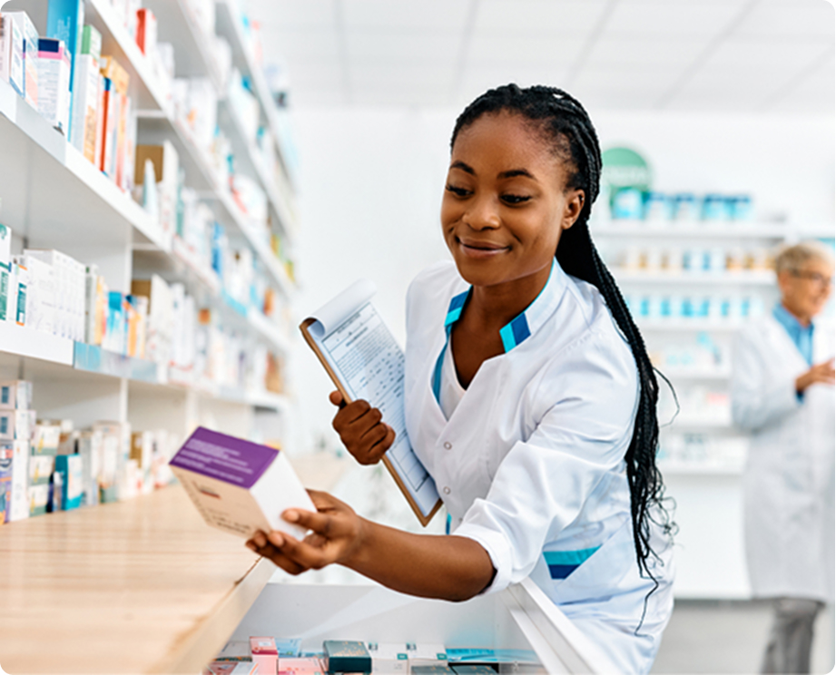 A pharmacist wearing a white coat holds a box of medication in a pharmacy. She has a clipboard and is smiling. In the background, another pharmacist is examining medicines on shelves. The setting is bright and organized.