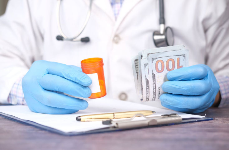 A doctor wearing blue gloves holds a pill bottle in one hand and a stack of hundred-dollar bills in the other, with a clipboard, papers, and pens on the table in front.