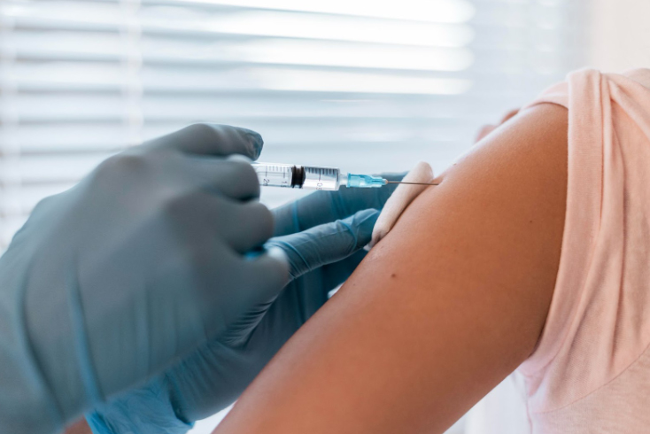 A close-up of a healthcare worker wearing blue gloves administering a vaccine injection into a person's upper arm. The person is wearing a light pink shirt and the background is softly blurred.