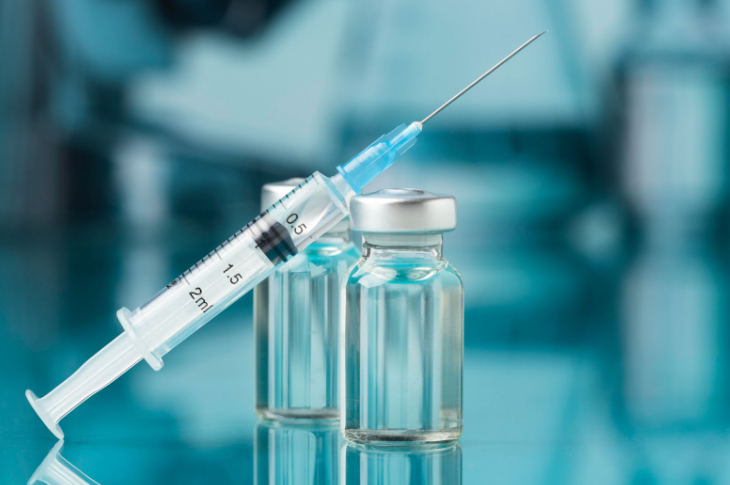 A syringe filled with liquid rests on two small glass vials containing a clear solution, set against a blue-toned, blurred medical background.