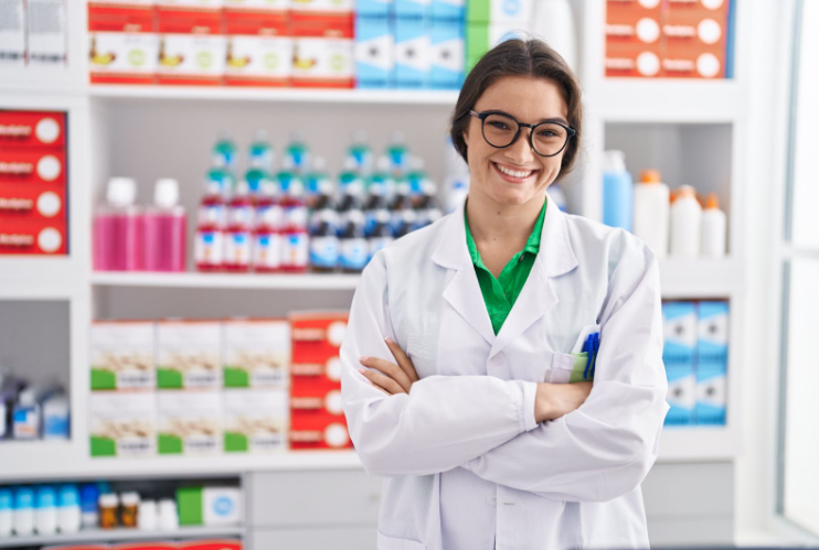 A smiling pharmacist wearing glasses and a white lab coat stands with arms crossed in a pharmacy, with shelves of medicine and colorful bottles in the background.