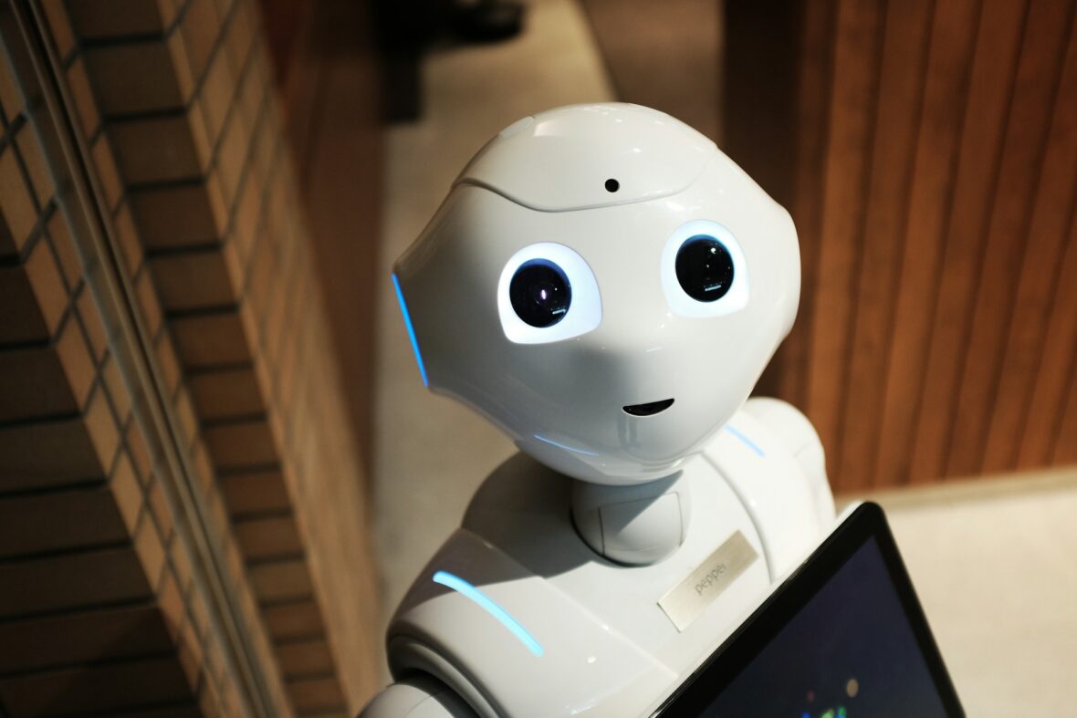 A white humanoid robot with large, expressive eyes and a tablet on its chest stands indoors, facing the camera in a well-lit setting with light wooden and brick walls.