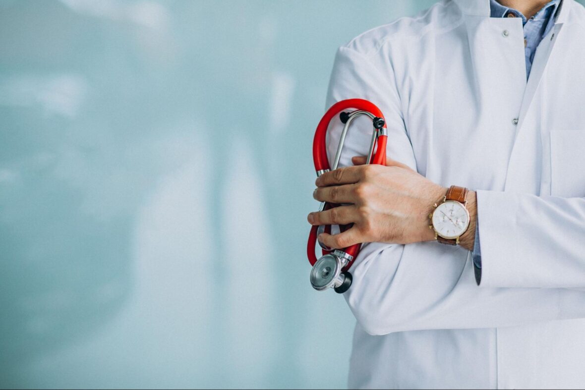 A person in a white lab coat holds a red stethoscope in one hand, with arms crossed. The face is not visible, and the background is a soft blue.